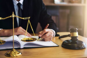 Avocat en costume noir signant un document juridique dans son cabinet, une balance de la justice et un marteau posés sur son bureau
