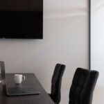 Bureau avec laptop ouvert, tasse blanche, deux chaises noires, écran mural, porte blanche