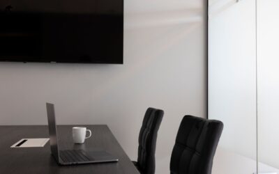 Bureau avec laptop ouvert, tasse blanche, deux chaises noires, écran mural, porte blanche